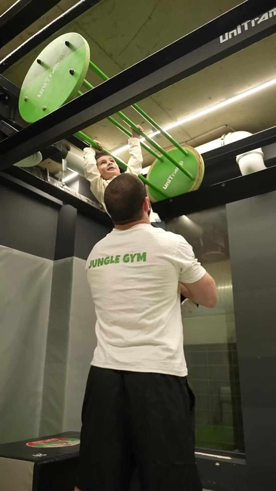 Gymnasia-ს JUNGLE GYM-ის მწვრთნელი ეხმარება პატარა ბავშვს მიწვდეს მწვანე საცოცი ღეროებამდე.